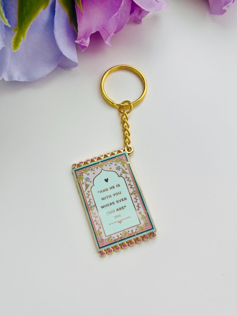 May include: Gold-colored keychain with a rectangular pendant. The pendant has a light blue background with the text "AND HE IS WITH YOU WHERE EVER YOU ARE". The edges are pink and gold, with floral accents.
