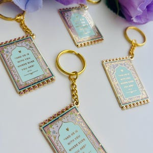 May include: Gold-toned keychains with rectangular pendants. Each pendant features a light blue background with the text "AND HE IS WITH YOU WHERE EVER YOU ARE." The borders are decorated with floral patterns and pink accents. The keychains are arranged on a white surface.