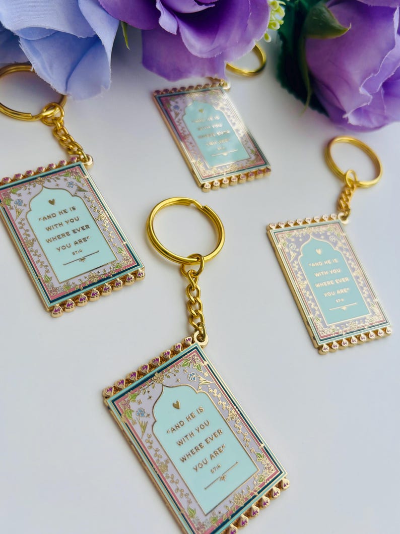 May include: Gold-toned keychains with rectangular pendants. Each pendant features a light blue background with the text "AND HE IS WITH YOU WHERE EVER YOU ARE." The borders are decorated with floral patterns and pink accents. The keychains are arranged on a white surface.