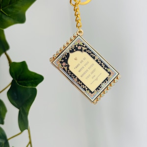 May include: Gold-toned keychain with a rectangular pendant featuring a floral design and the text "AND HE IS WITH YOU WHERE EVER YOU ARE". The pendant has a gold border with small crystals.