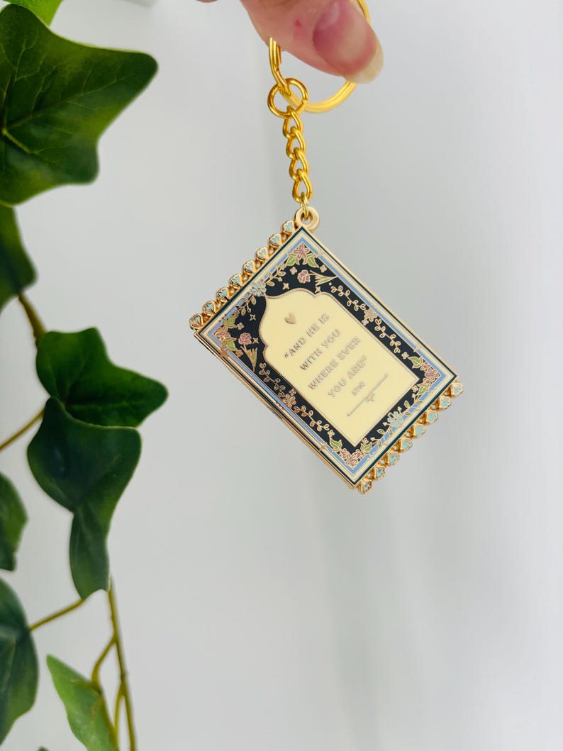 May include: Gold-toned keychain with a rectangular pendant featuring a floral design and the text "AND HE IS WITH YOU WHERE EVER YOU ARE". The pendant has a gold border with small crystals.