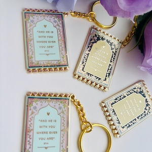 May include: Four rectangular keychains with gold-colored chains and rings. Each keychain has a decorative border and features the text "AND HE IS WITH YOU WHERE EVER YOU ARE". The keychains have a light blue, pink, and gold color scheme.