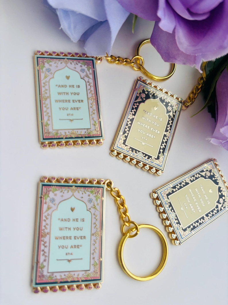 May include: Four rectangular keychains with gold-colored chains and rings. Each keychain has a decorative border and features the text "AND HE IS WITH YOU WHERE EVER YOU ARE". The keychains have a light blue, pink, and gold color scheme.