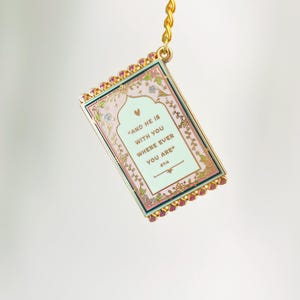 May include: A rectangular keychain with a gold chain and clasp. The keychain has a pink and green background with the text "AND HE IS WITH YOU WHERE EVER YOU ARE" and floral accents. The edges are trimmed with pink jewels.