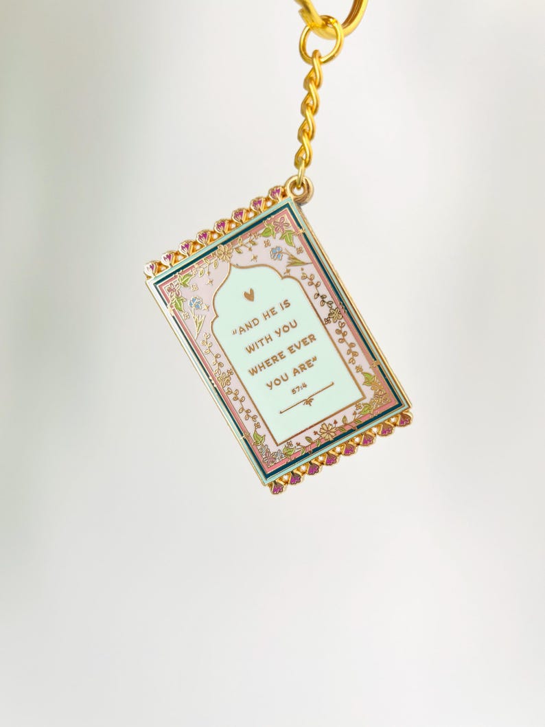 May include: A rectangular keychain with a gold chain and clasp. The keychain has a pink and green background with the text "AND HE IS WITH YOU WHERE EVER YOU ARE" and floral accents. The edges are trimmed with pink jewels.