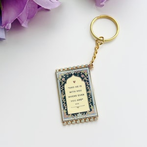 May include: A gold-colored keychain with a rectangular pendant. The pendant has a cream-colored background with the text "AND HE IS WITH YOU WHERE EVER YOU ARE" and floral border. The keychain has a gold ring and chain.