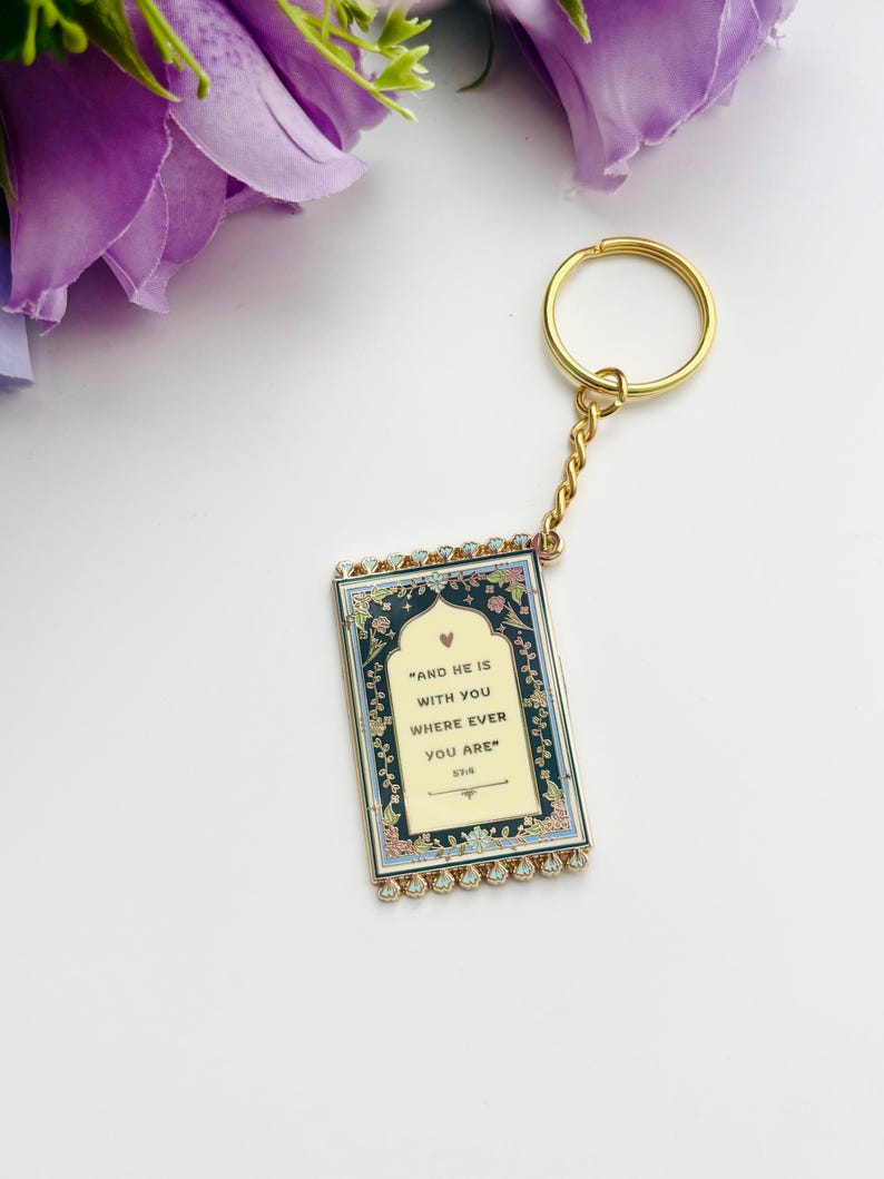 May include: A gold-colored keychain with a rectangular pendant. The pendant has a cream-colored background with the text "AND HE IS WITH YOU WHERE EVER YOU ARE" and floral border. The keychain has a gold ring and chain.