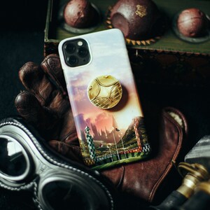 May include: A phone case with a golden snitch and a Hogwarts castle scene. The case is on a brown leather glove with a pair of goggles next to it.
