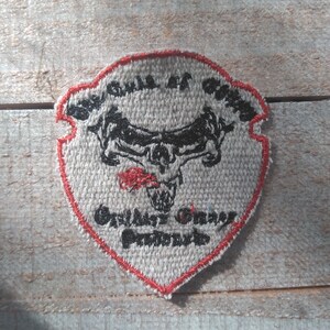 May include: A shield-shaped patch with a gray background and a red border. The patch features a black skull with horns and the text "Gulls of Gotham" and "Gallery Owner Presents". The patch is likely for a biker or motorcycle club.
