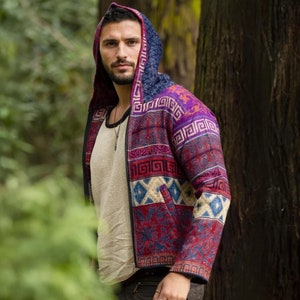 Puede incluir: Un hombre con una chaqueta con capucha de estampado colorido con cierre de cremallera. La chaqueta tiene un diseño geométrico con tonos de rojo, morado, azul y blanco. El hombre está de pie en un entorno boscoso.
