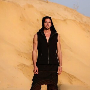 May include: A man wearing a black hooded vest and brown pants stands in a desert landscape. The vest has a zipper and a hood. The man is looking at the camera.