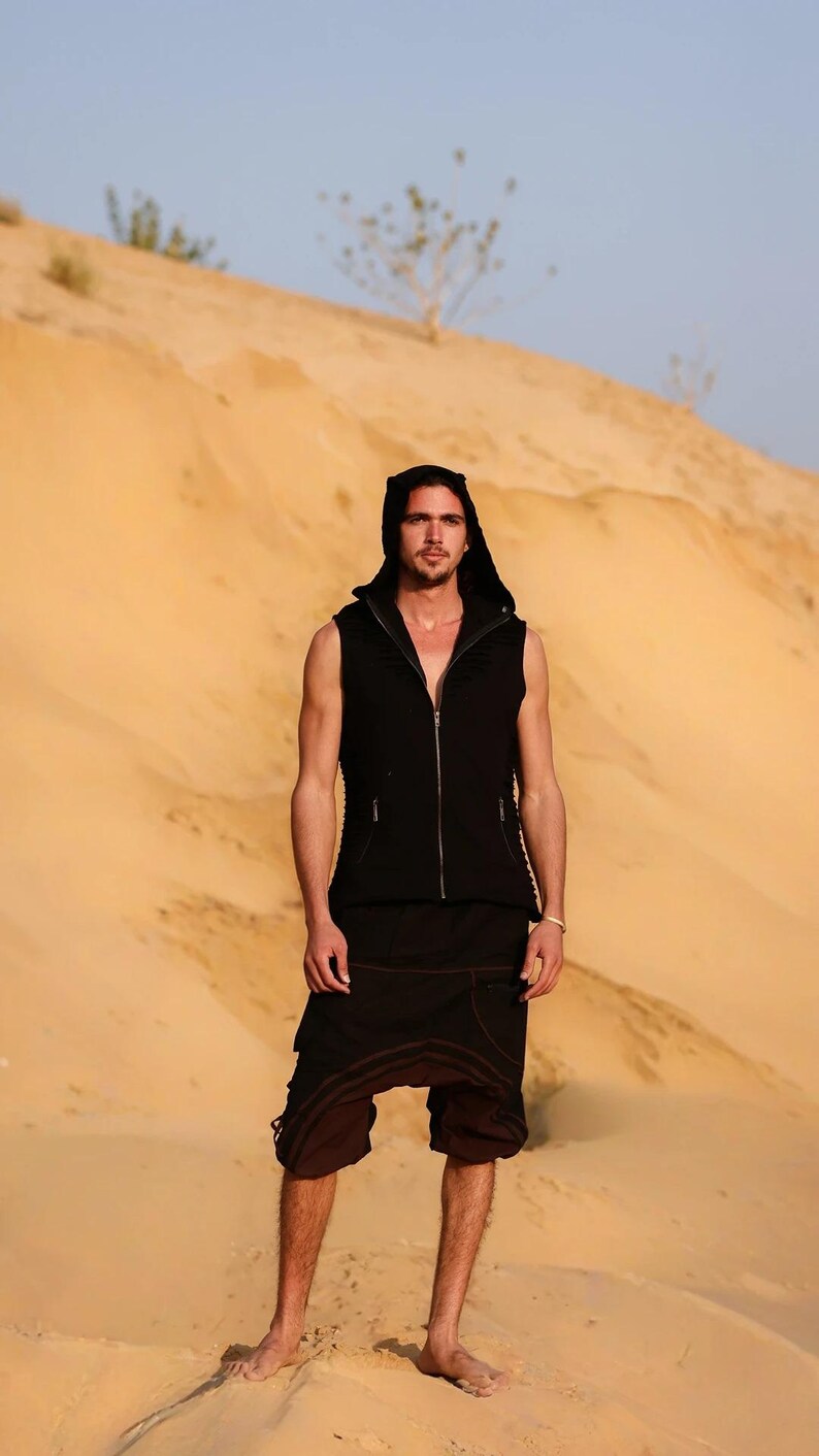 May include: A man wearing a black hooded vest and brown pants stands in a desert landscape. The vest has a zipper and a hood. The man is looking at the camera.