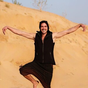 May include: A person wearing a black sleeveless vest with a hood, black pants, and a black hooded top. The person is standing in a desert with sand dunes in the background.