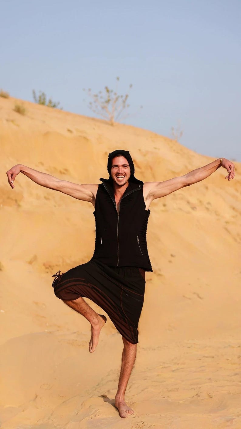 May include: A person wearing a black sleeveless vest with a hood, black pants, and a black hooded top. The person is standing in a desert with sand dunes in the background.