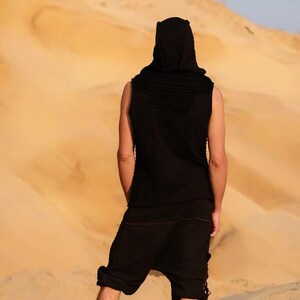 May include: A person wearing a black sleeveless hooded top and black pants stands in a desert landscape. The person is facing away from the camera and looking out at the horizon.