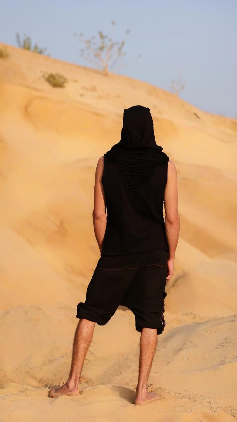 May include: A person wearing a black sleeveless hooded top and black pants stands in a desert landscape. The person is facing away from the camera and looking out at the horizon.
