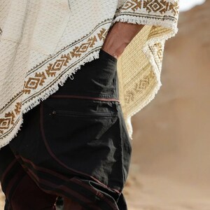 May include: A person wearing a white poncho with a brown and green geometric pattern and black pants with a brown stripe.