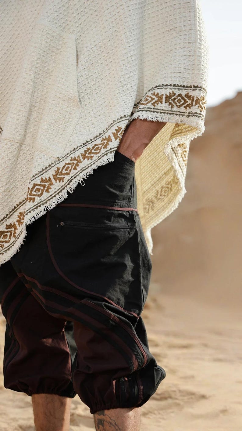 May include: A person wearing a white poncho with a brown and green geometric pattern and black pants with a brown stripe.