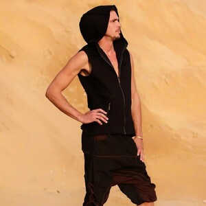 May include: A man wearing a black hooded vest and brown pants stands in a desert landscape. The vest has a zipper and a hood. The pants have a drawstring waist and are loose fitting.