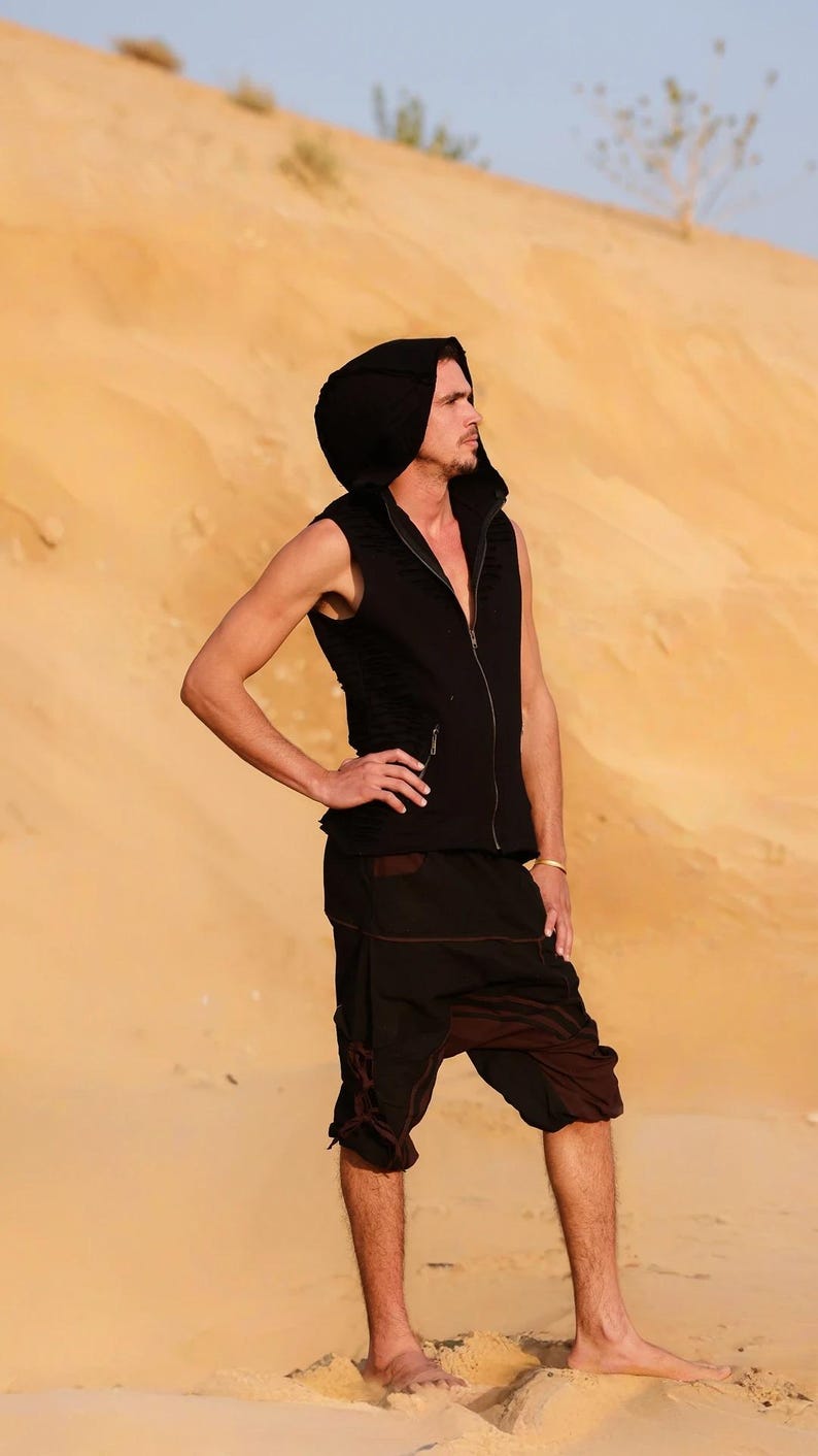 May include: A man wearing a black hooded vest and brown pants stands in a desert landscape. The vest has a zipper and a hood. The pants have a drawstring waist and are loose fitting.