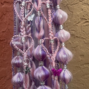 Puede incluir: Extensiones de cabello trenzadas en tonos morados y rosas, adornadas con detalles plateados y abalorios. Las extensiones presentan trenzas en forma de corazón, cuentas decorativas y abalorios de mariposa, creando un aspecto caprichoso y decorativo.