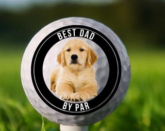 May include: A golf ball on a tee with a circular graphic featuring a golden retriever puppy and the text "BEST DAD BY PAR". The golf ball is set against a backdrop of green grass and a blurred background.