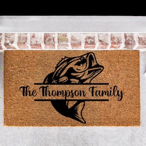 May include: A brown coir doormat with a black silhouette of a bass fish jumping out of the water. The text "The Thompson Family" is printed in black on a white banner below the fish.