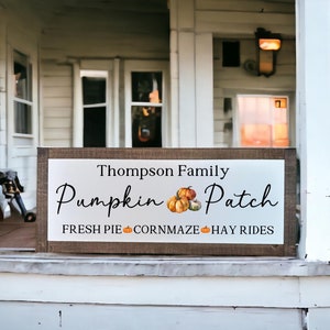 May include: A white sign with brown wood trim on a white porch. The sign reads "Thompson Family Pumpkin Patch" with a small pumpkin graphic and the text "Fresh Pie, Corn Maze, Hay Rides".