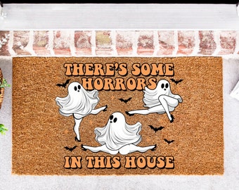 May include: A rectangular coir doormat with the text "THERE'S SOME HORRORS IN THIS HOUSE" in orange lettering. Three cartoon ghosts in various poses are featured, along with black bat silhouettes. The mat is brown.