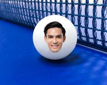 May include: A white ping pong ball with a man's face printed on it. The ball is on a blue ping pong table.