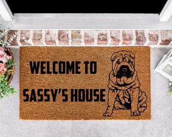 May include: A brown coir doormat with the text "Welcome to Sassy's House" in black lettering. A black and white illustration of a Shar-Pei dog is on the right side of the mat.
