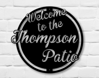 May include: Black metal sign with the words "Welcome to the Thompson Patio" in a decorative font. The sign is circular with a silver outline and is mounted on a white brick wall. Green bushes are in the foreground.