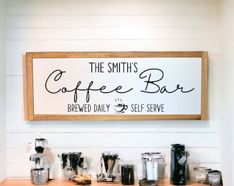 May include: A rectangular wooden sign with the text "The Smith's Coffee Bar" in black script. Below, it reads "Brewed Daily Self Serve" with a coffee cup icon. The sign is mounted on a white paneled wall.