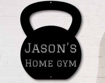 May include: Black metal kettlebell shaped sign with the text "JASON'S HOME GYM" in white lettering. The sign is mounted on a white brick wall.
