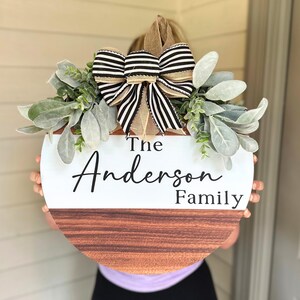 May include: A round wooden door hanger with a white top half and a brown wood bottom half. The top half has the text "The Anderson Family" in black script font. The hanger is decorated with a black and white striped bow and greenery.