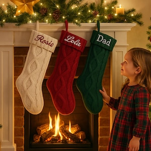 May include: Three cable-knit Christmas stockings in cream, red, and green hang above a fireplace. Each stocking is personalized with a name: "Rosie", "Lola", and "Dad". A child in a plaid dress looks at the stockings.