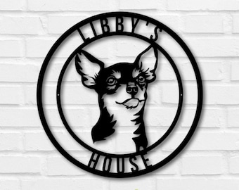 May include: Black metal sign featuring a Chihuahua dog's head inside a circular frame. The words "LIBBY'S" and "HOUSE" are arched above and below the dog's image. The sign is mounted on a white brick wall, with green foliage in the foreground.