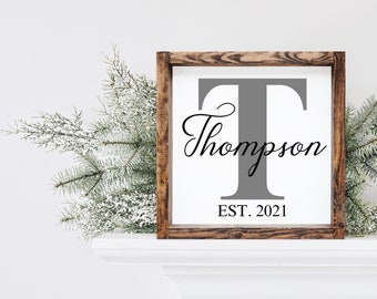 May include: A wooden sign with a gray letter T and the text "Thompson EST. 2021" in black. The sign is on a white mantel with a green pine branch to the left.