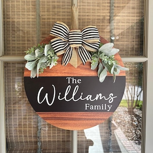 May include: A round wooden door hanger with a black and white striped bow, green foliage, and the text "The Williams Family".