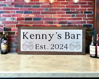 May include: A brown wooden sign with a silver metal insert. The sign reads "Kenny's Bar Est. 2024" with two black and white illustrations of beer mugs.