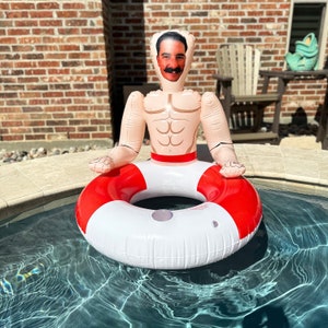 May include: Inflatable pool float shaped like a man with a face, muscular torso, and arms. The float is seated in a red and white life preserver ring, floating in a pool. The man's face has a mustache and dark hair.
