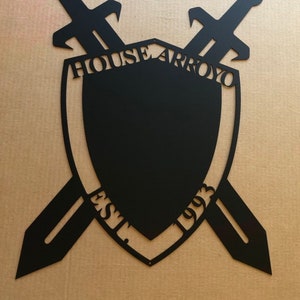 May include: Black metal shield with two crossed swords, the shield has the text "HOUSE ARROYO EST. 1993" around the edge.