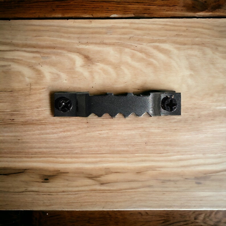 May include: A black metal picture hanging hook with two screws. The hook is attached to a light wood surface.