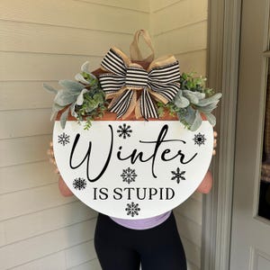 May include: A round, white wooden sign with the words "Winter IS STUPID" in black script and snowflake accents. The sign is decorated with greenery and a striped bow.