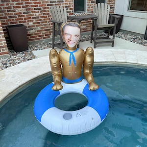 May include: An inflatable pool float shaped like a shirtless man with a smiling face. The float is gold and blue, with a blue and white inner tube. It is floating in a pool of water.