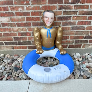 May include: An inflatable pool float shaped like a torso with a face, gold arms and torso, and a blue and white inner tube. The face is a photo of a person. The float is sitting on a bed of rocks in front of a brick wall.