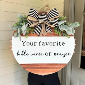 May include: A round wooden door hanger with a white background and black text that reads "Your favorite bible verse or prayer". The hanger is decorated with a black and white striped bow and green foliage.