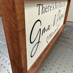 May include: A wooden sign with a white background and black lettering. The sign reads "There's No Place Like Grandma & Papa's House". The sign is framed in a dark brown wood frame.