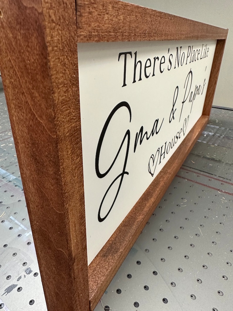 May include: A wooden sign with a white background and black lettering. The sign reads "There's No Place Like Grandma & Papa's House". The sign is framed in a dark brown wood frame.