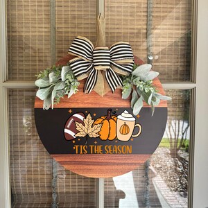 May include: A round wooden fall-themed wreath with a black and brown striped bow and greenery. The wreath features a football, pumpkins, a leaf, and a pumpkin spice latte graphic. The text "'Tis the Season" is printed on the bottom.